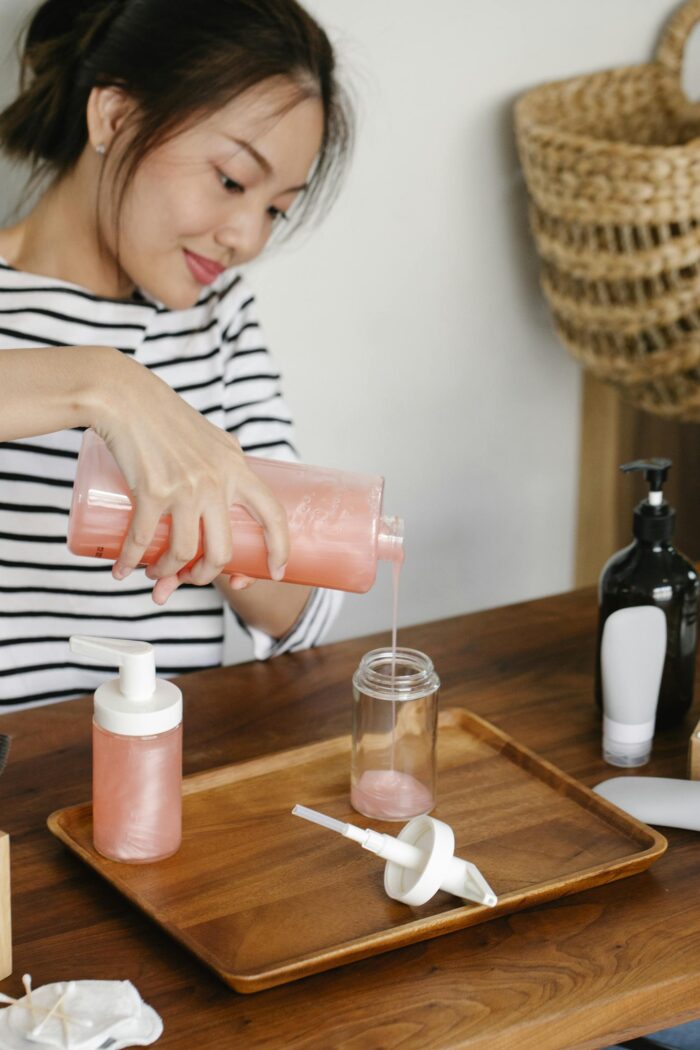 Crop Asian woman in casual clothes pouring peach coloured soap from opened soap dispenser into plastic bottle on tray while sitting at table