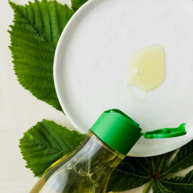 Top view of open bottle of cosmetic oil placed flat on table near white plate with small amount of oil on it on big fresh green leaf