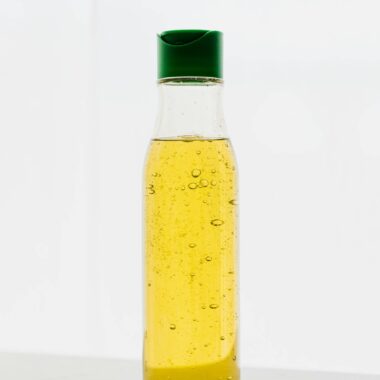 Yellow body oil with bubbles in transparent plastic bottle with green cap on table against white background